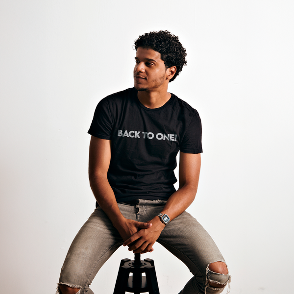 Male model wearing a black 100% cotton t-shirt from Beard and Hat Actor with the phrase “BACK TO ONE!” printed in light gray, seated on a black stool against a white background