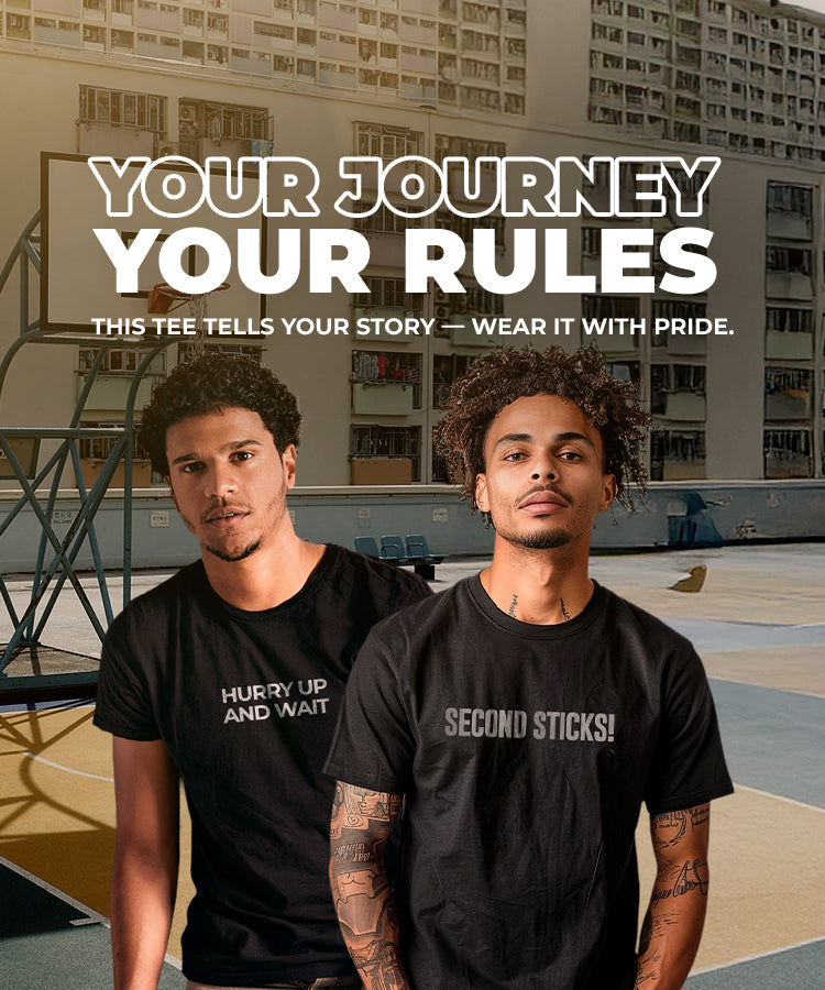 Two male models wearing black 100% cotton t-shirts from Beard and Hat Actor—one with the phrase “HURRY UP AND WAIT” and the other with “SECOND STICKS” printed in light gray. They stand on an urban basketball court with high-rise buildings in the background. Overlay text reads: “YOUR JOURNEY, YOUR RULES — This tee tells your story, wear it with pride.”