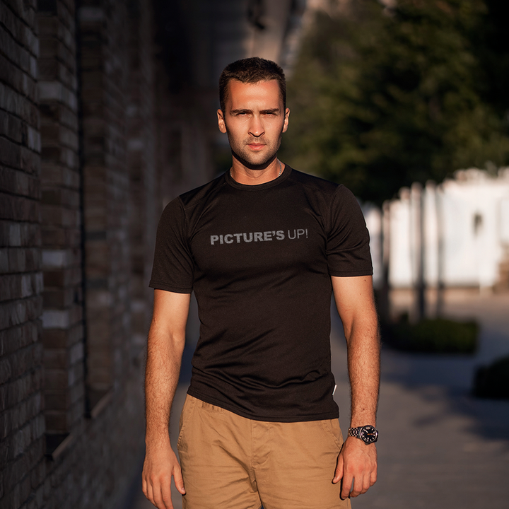Male model wearing a black 100% cotton t-shirt from Beard and Hat Actor with the phrase “PICTURE’S UP!” printed in light gray