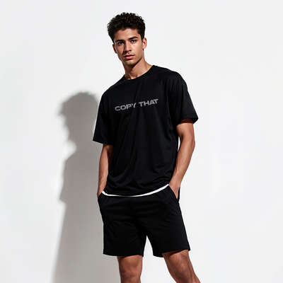 Male model wearing a black 100% cotton t-shirt from Beard and Hat Actor with the phrase “COPY THAT” printed in light gray across the chest, standing against a white wall with hands in pockets