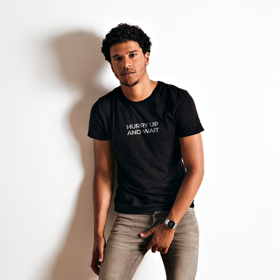 Male model wearing a black 100% cotton t-shirt from Beard and Hat Actor with the phrase “HURRY UP AND WAIT” printed in light gray across the chest, standing against a white wall with relaxed posture
