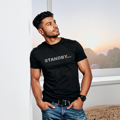 Male model wearing a black 100% cotton t-shirt from Beard and Hat Actor with the word “STANDBY…” printed in light gray across the chest, standing by a window at sunset
