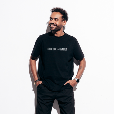 Male model wearing a black 100% cotton t-shirt from Beard and Hat Actor with the phrase “CHECK THE GATE” printed in light gray across the chest, standing against a white background with hands in pockets and smiling