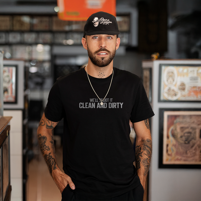 Male model wearing a black 100% cotton t-shirt from Beard and Hat Actor with the phrase “WE’LL SHOOT IT CLEAN AND DIRTY” printed in light gray across the chest. He is also wearing a black Beard and Hat Actor baseball cap and standing in a tattoo studio