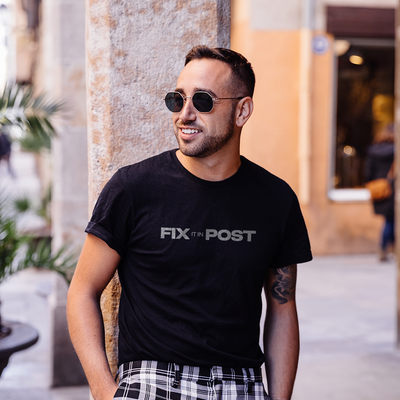 Male model wearing a black 100% cotton t-shirt from Beard and Hat Actor with the phrase “FIX IT IN POST” printed in light gray across the chest, leaning casually against a column in an urban outdoor setting