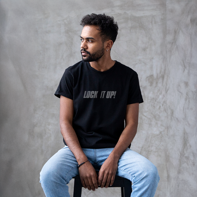 Male model wearing a black 100% cotton t-shirt from Beard and Hat Actor with the phrase “LOCK IT UP!” printed in light gray across the chest, seated on a stool against a textured gray backdrop