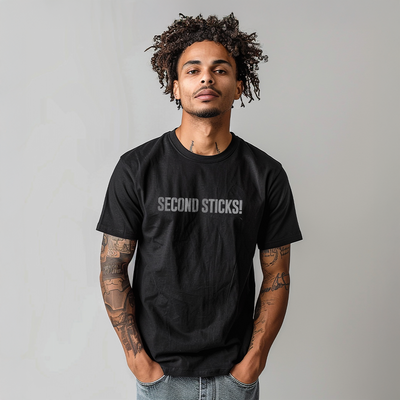 Male model wearing a black 100% cotton t-shirt from Beard and Hat Actor with the phrase “SECOND STICKS!” printed in light gray across the chest, standing against a neutral gray background