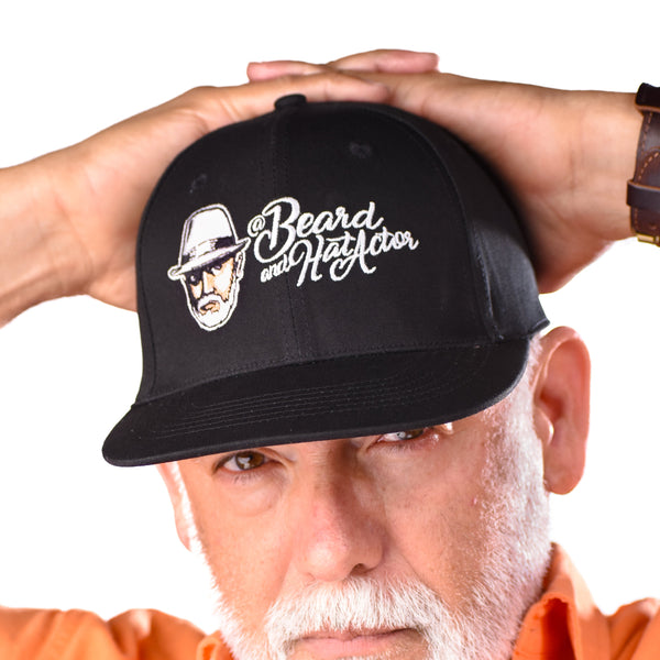 Male model wearing a black flat bill fitted cap with the Beard and Hat Actor logo embroidered on the front, shown close-up with a serious expression and an orange shirt
