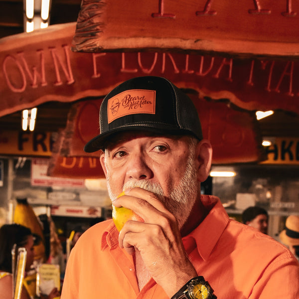 Older male model wearing a black and gray 6-panel structured trucker cap with a curved bill and a leather patch featuring the Beard and Hat Actor logo. He is dressed in an orange shirt, holding a mango near his mouth inside a colorful fruit market with 