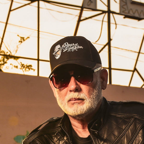 Older male with a white beard wearing a black 6-panel structured curved bill cap with the Beard and Hat Actor logo embroidered on the front. He is seated on a motorcycle, wearing a black leather jacket and sunglasses, in front of a sunset-lit industrial background