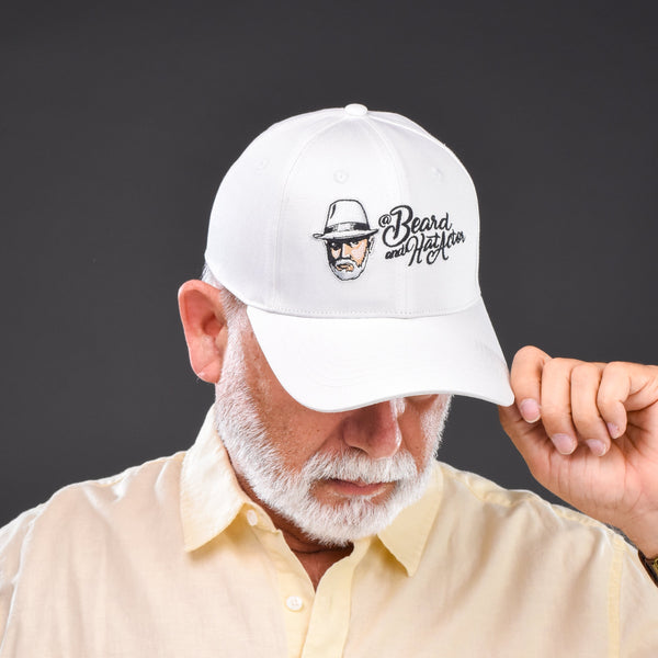 Older male model wearing a white 6-panel structured curved bill cap with the Beard and Hat Actor logo embroidered on the front. He is dressed in a light yellow shirt, looking down while slightly lifting the brim of the cap with one hand